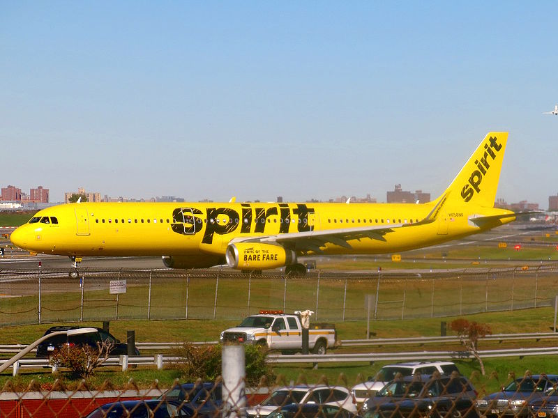 A Spirit Airlines Airbus A321 in the newest livery is taxiing from Taxiway B to Runway 4 for takeoff clearance, as photographed from Planeview Park. Runway 4/22 runs behind the aircraft, and the Grand Central Parkway is visible in the bottom of the picture.
